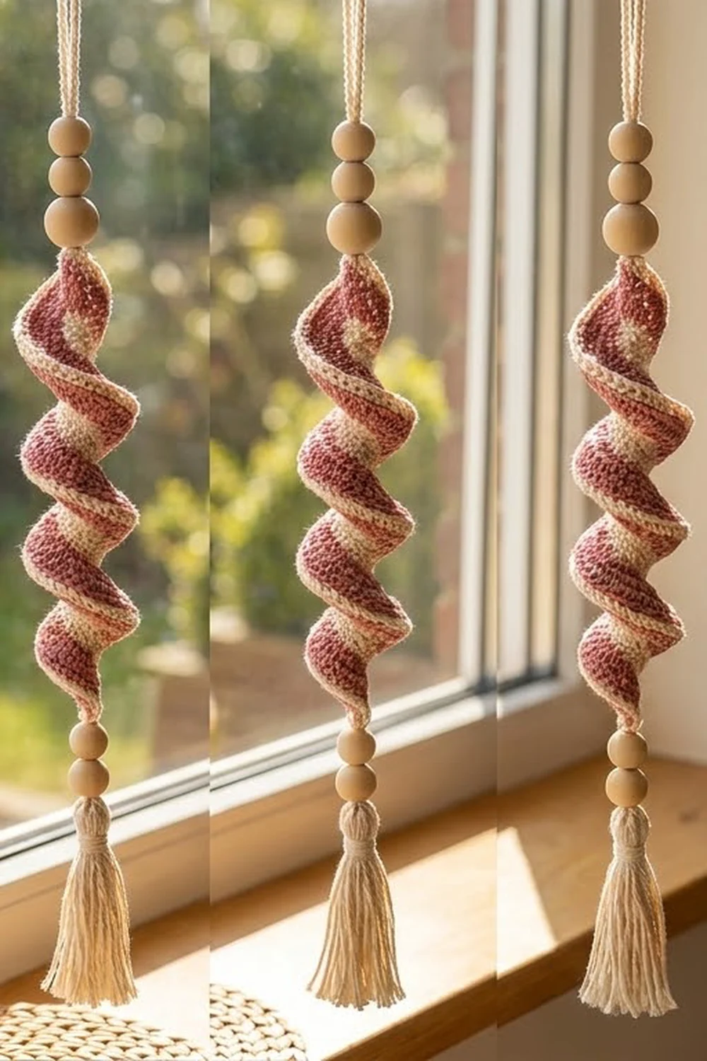 Three handmade window ornaments with wooden beads and burgundy knitted fabric spirals hanging by twine in sunlight.