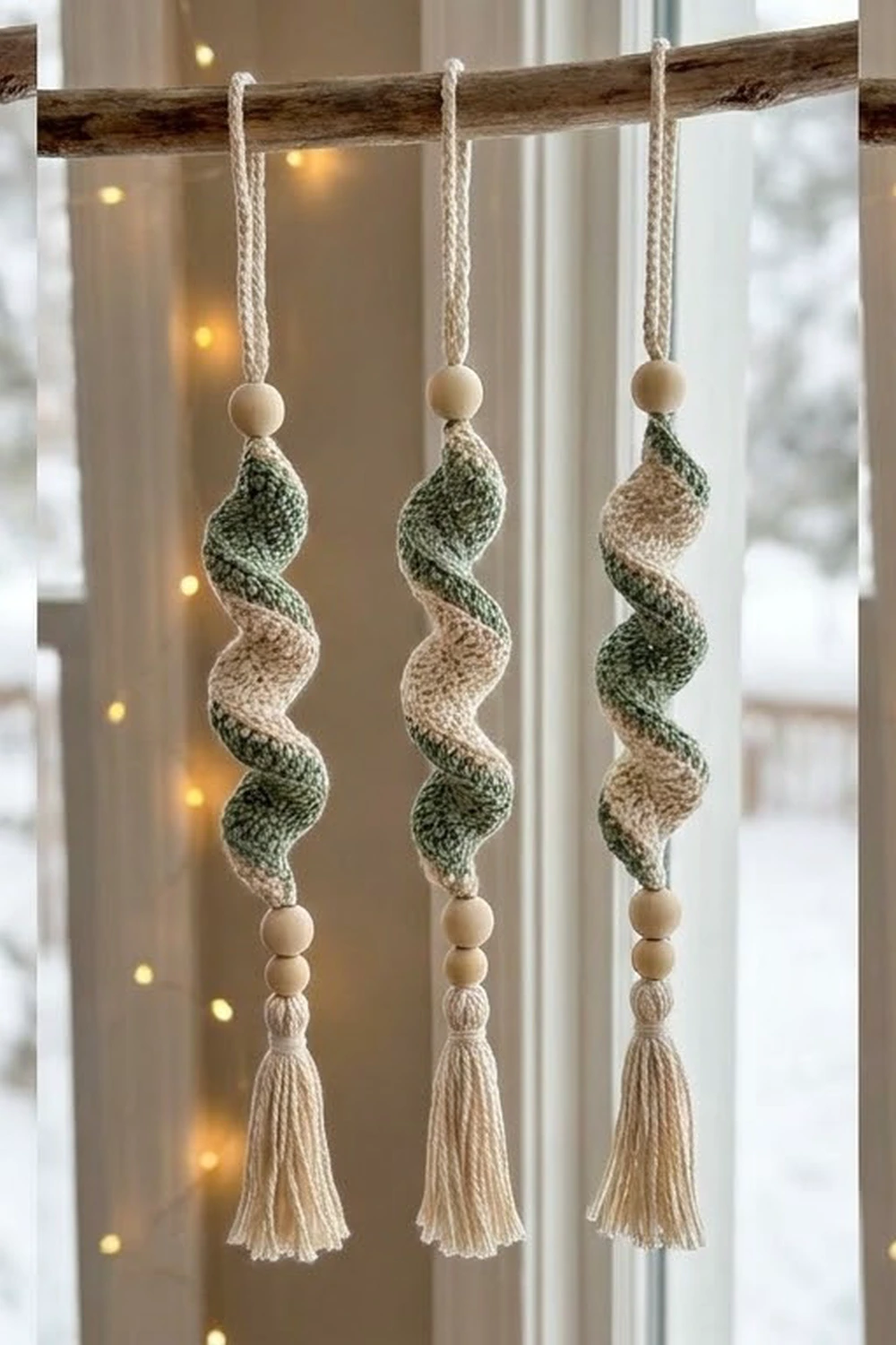 Three knitted spiral ornaments with wooden beads and tassel details hang from a rustic branch in a window.