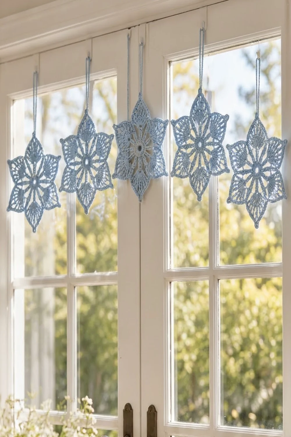 Delicate blue-gray crocheted snowflakes hang from strings in a sunlit window overlooking trees.