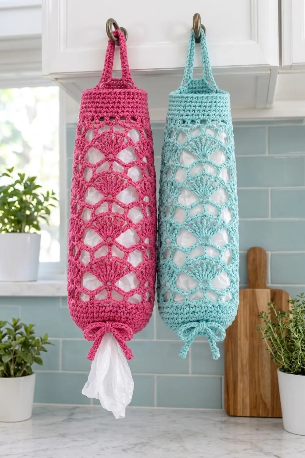Hot pink and mint crocheted bag dispensers with lace patterns hang from kitchen cabinet beside green potted herbs.