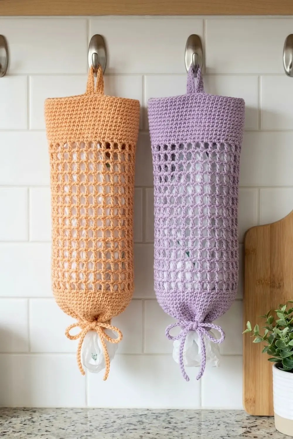 Two crocheted mesh produce bags in orange and purple hanging from metal hooks on a white kitchen tile backsplash.