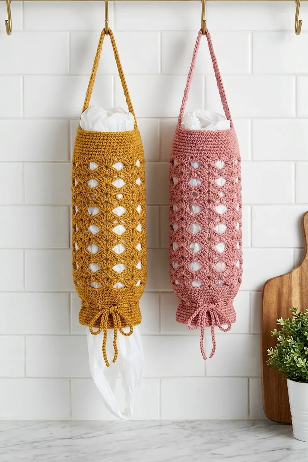 Mustard and mauve crocheted tissue holders with openwork diamond designs hanging from brass hooks on white subway tile.