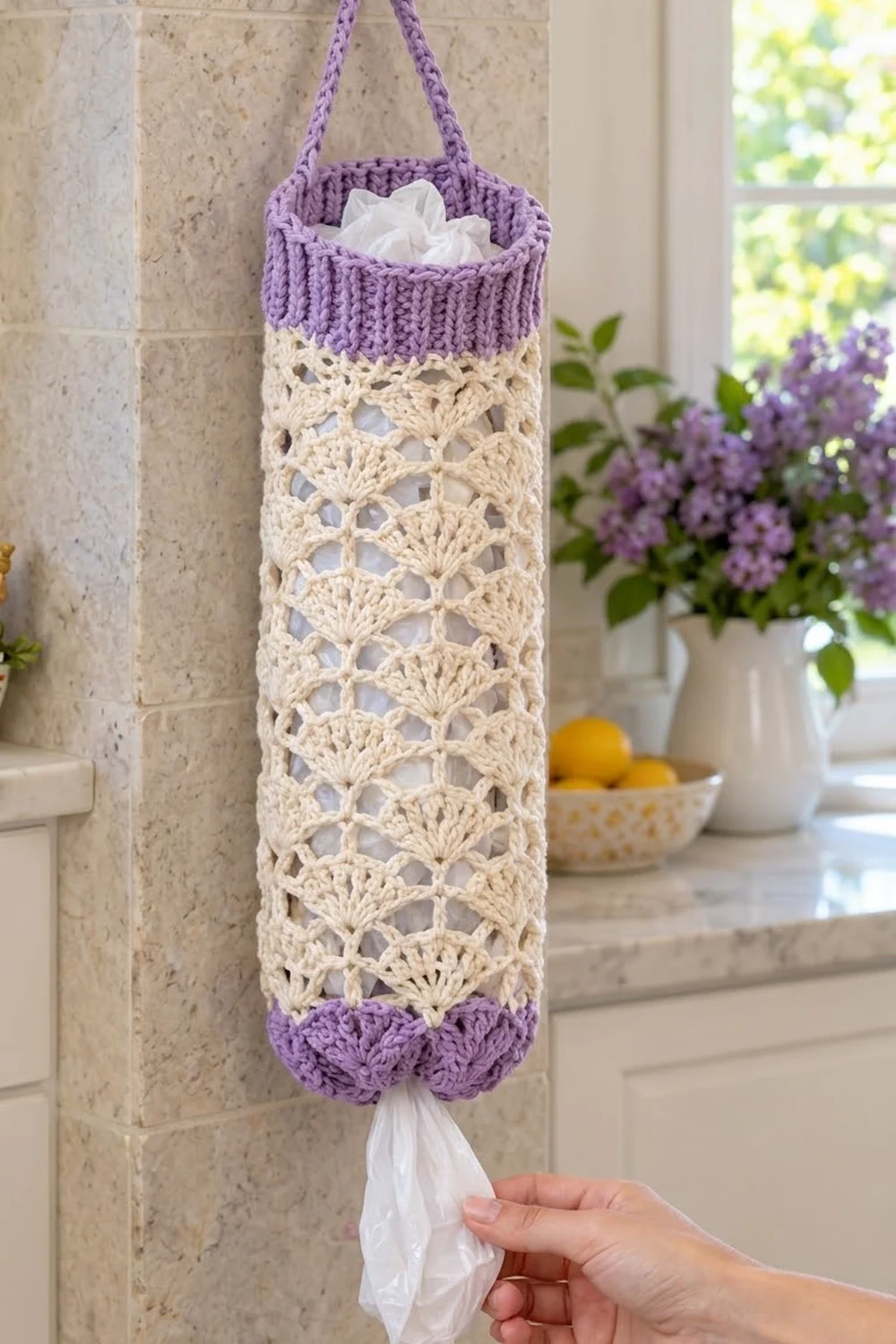 Lavender and cream crochet tissue holder with shell stitch pattern hanging on kitchen tile wall near purple flowers.