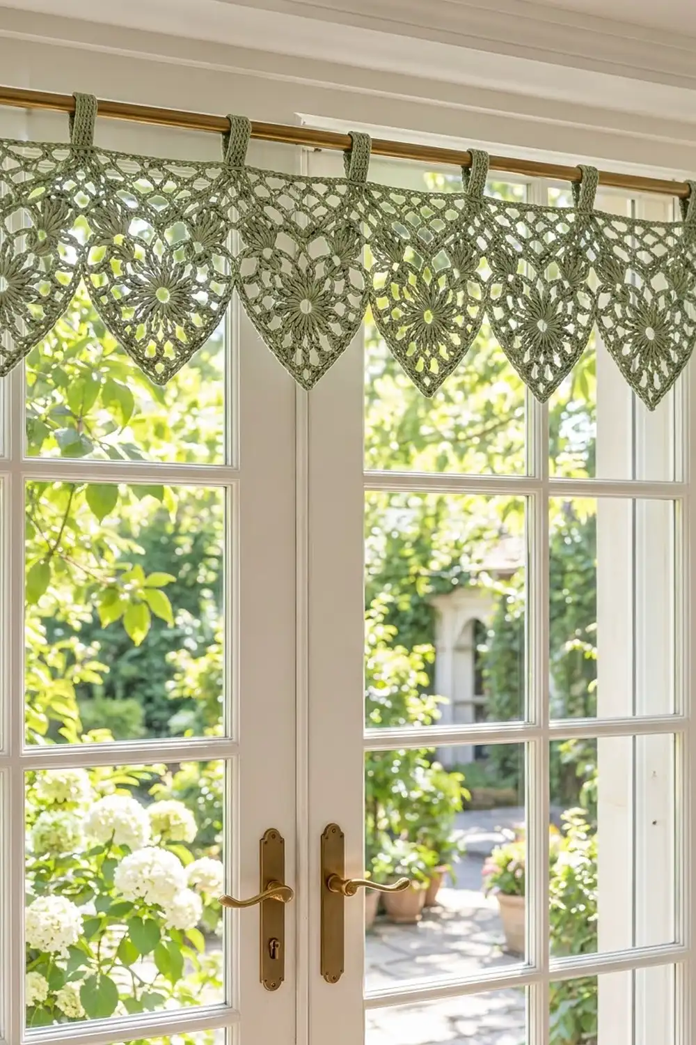 Crocheted sage-green valance with floral motifs hanging above a window framing a sunlit garden with white flowering shrubs.