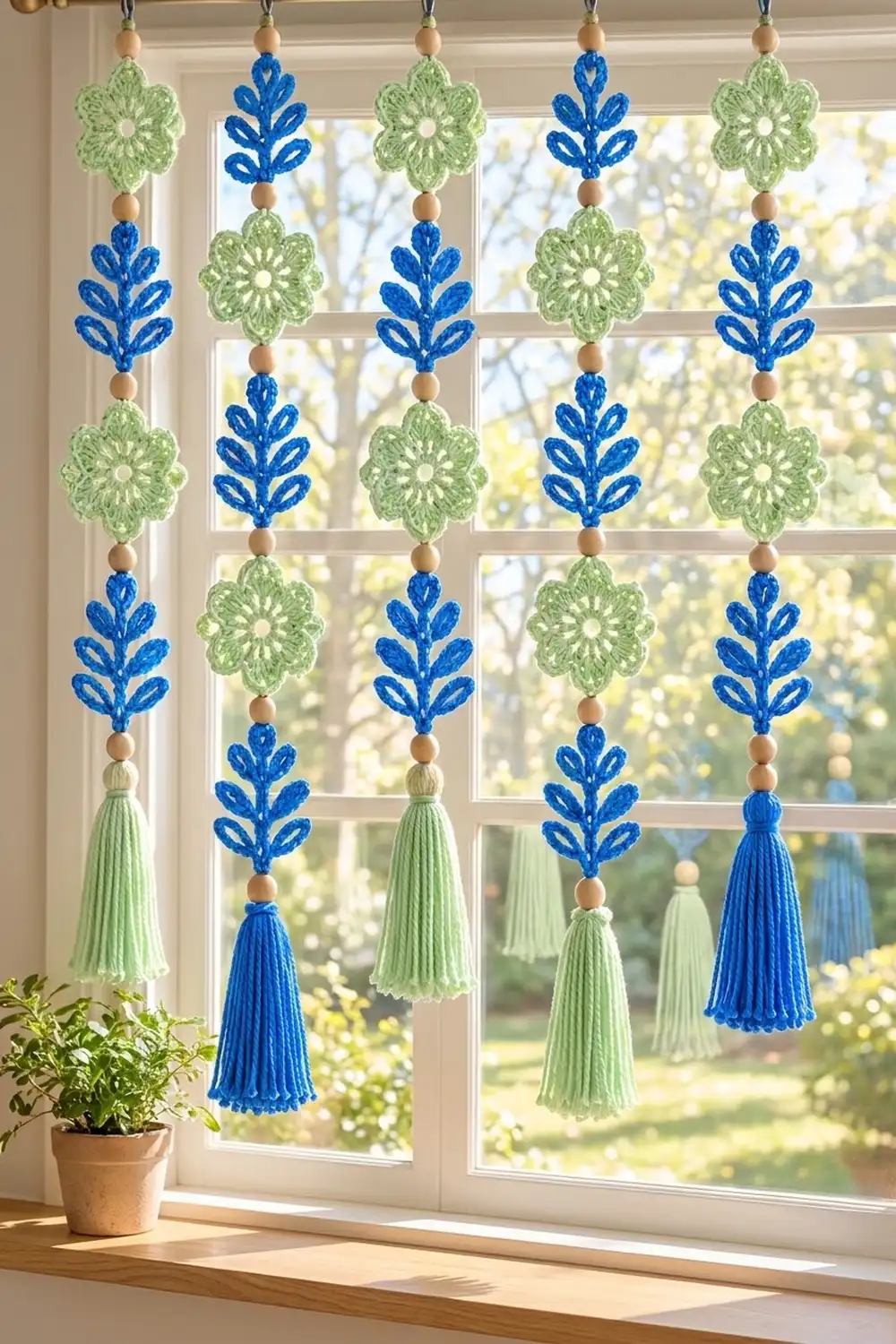 Crochet window garland with alternating green flowers and blue leaves with wooden beads and tassels, hanging in a sunlit window.