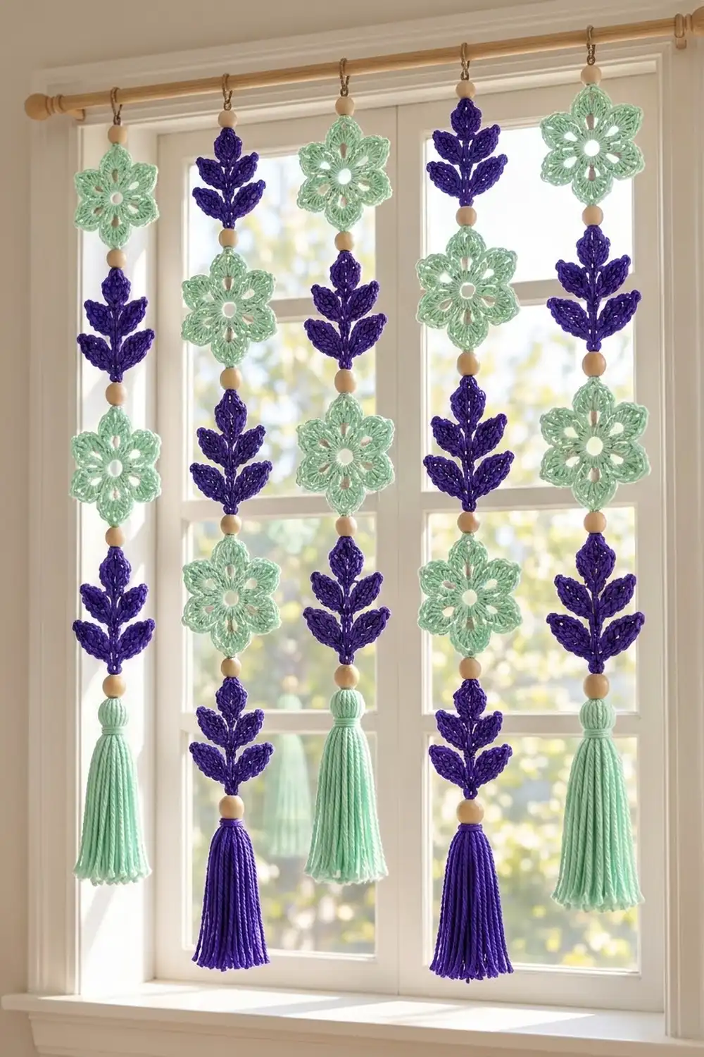 Navy and mint crochet flower garlands with wooden beads and tassels hang from a wooden rod in a bright window.
