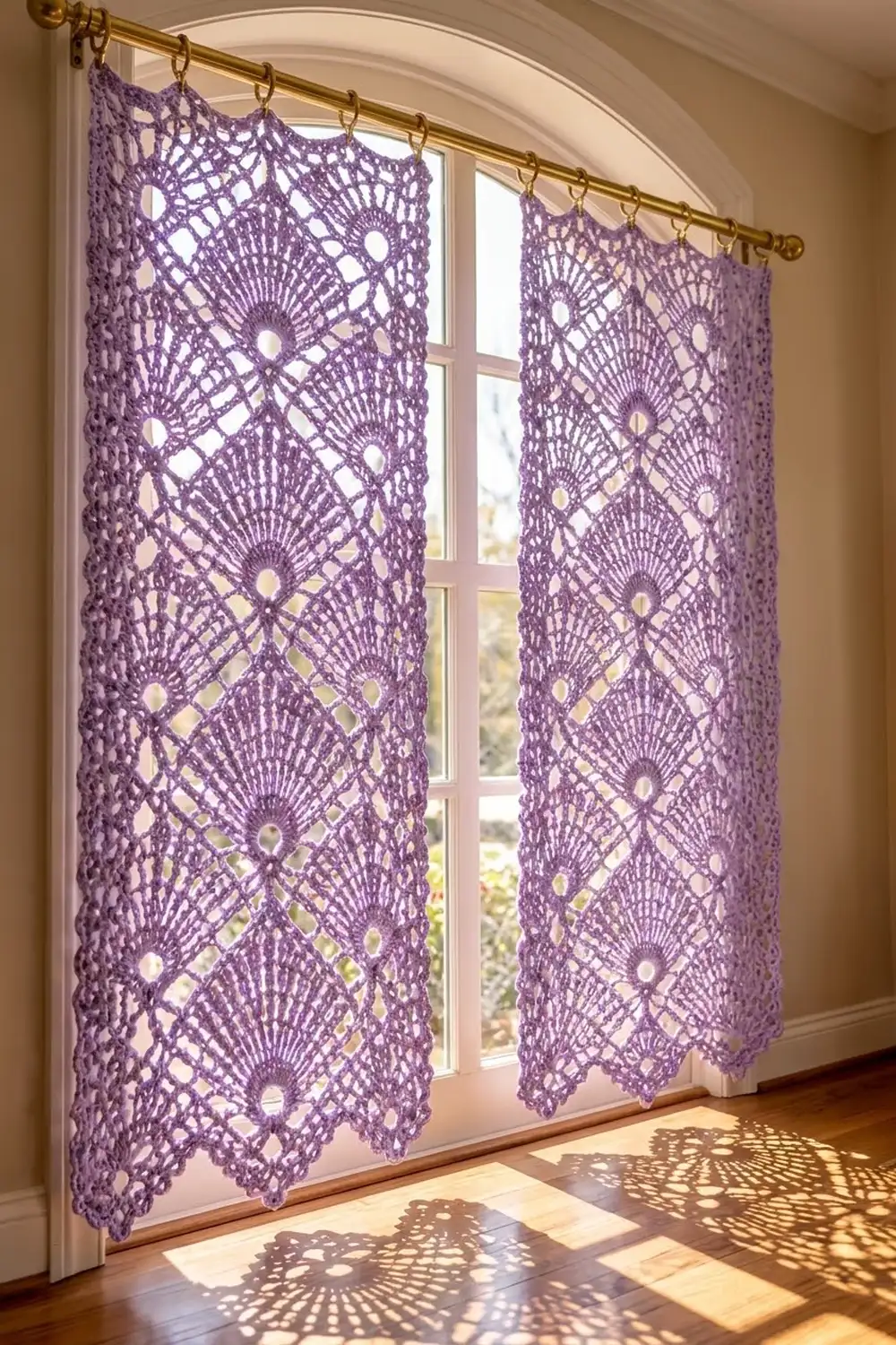 Purple crocheted lace curtains with geometric fan motifs hang on a brass rod, casting delicate shadows through a sunny window.