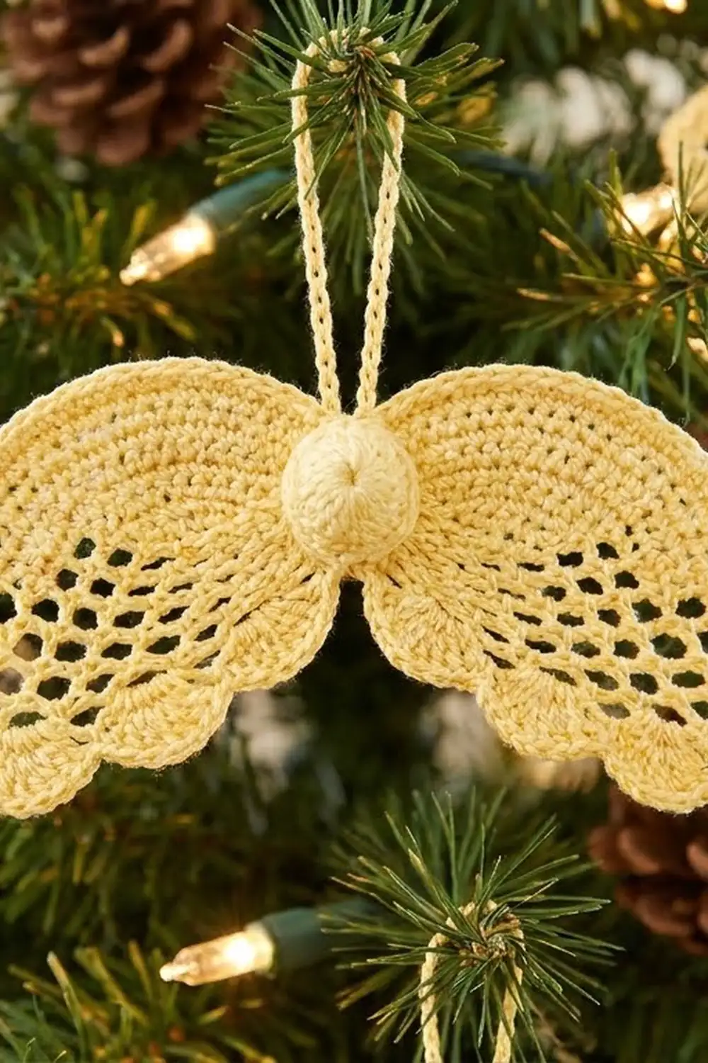 Pale yellow crocheted angel wings ornament with openwork pattern hanging on fresh evergreen boughs.