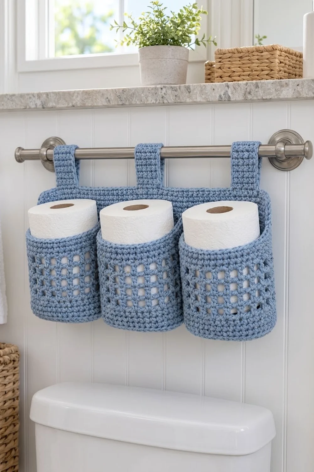Pale blue crocheted toilet paper holder with three compartments holding white rolls, hanging on chrome rod in bright bathroom.
