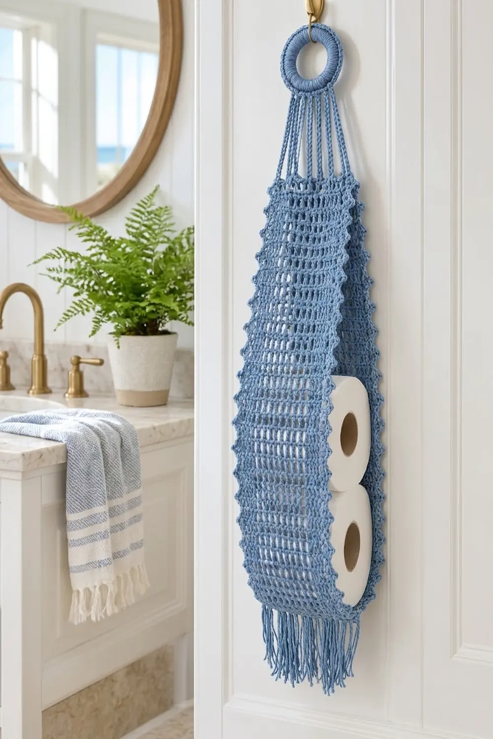 Blue crocheted toilet paper holder with macramé design and fringe hanging in a bright bathroom.