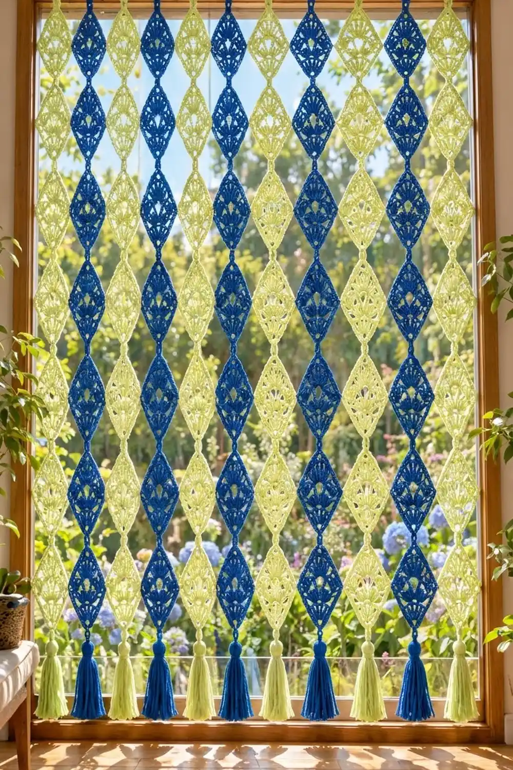 Navy and pale-green crocheted window panel with diamond motifs and tassels frames a sunny garden view with blooming flowers.
