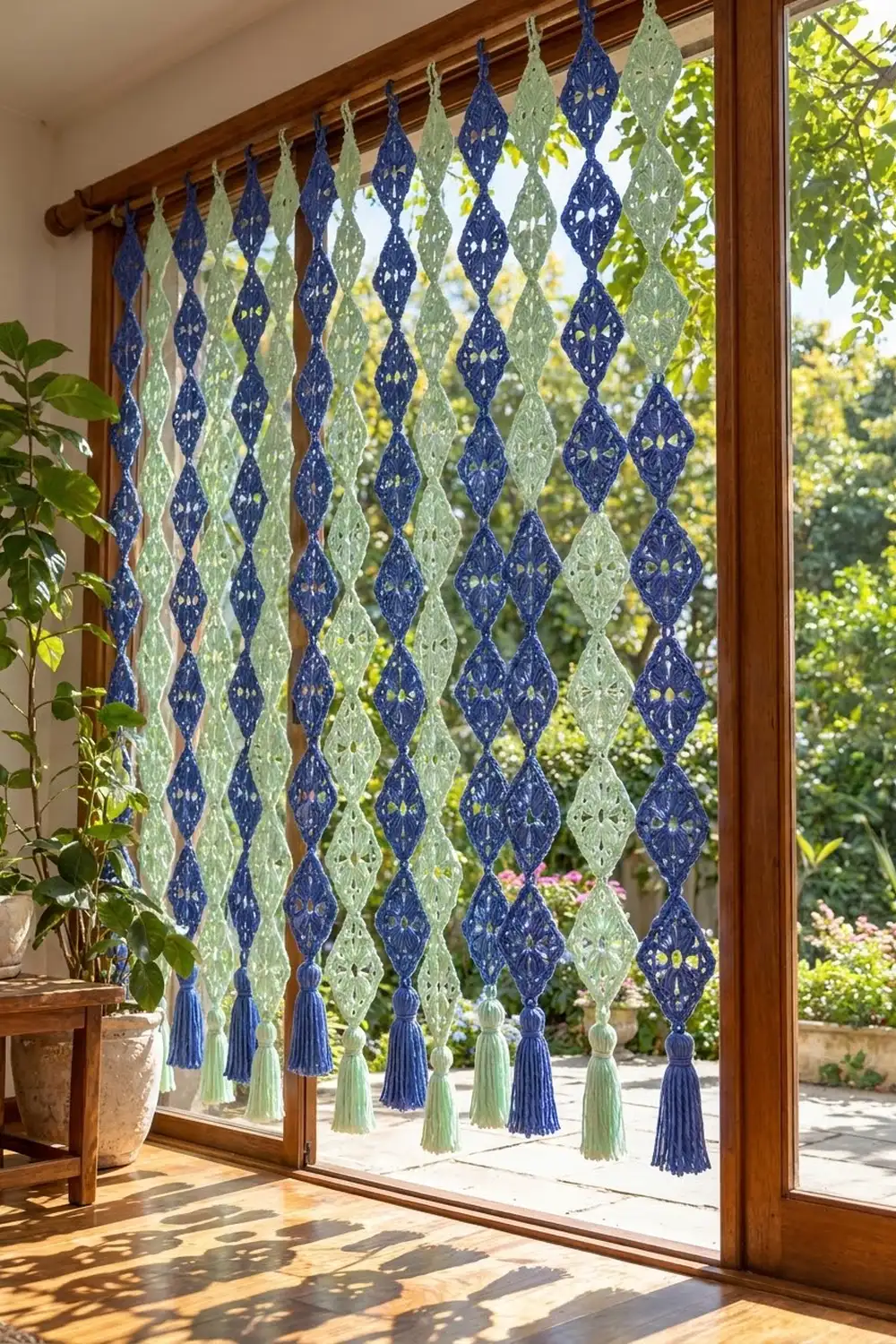 Crocheted blue and sage green diamond-patterned curtain with tassels hanging in a sunlit doorway overlooking a garden.