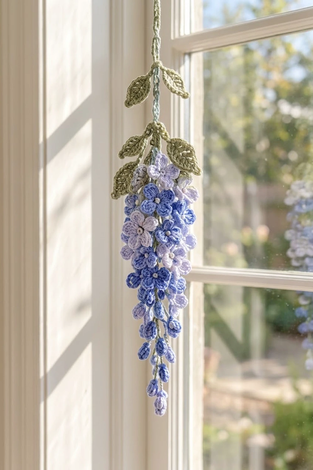 Crocheted blue and purple wisteria with green leaves hangs in a sunlit window overlooking a garden.