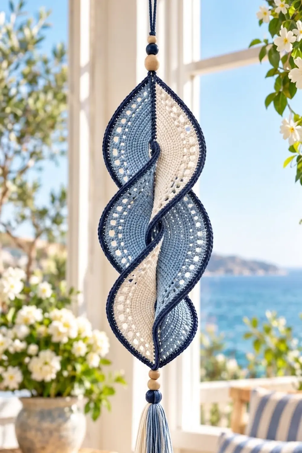 Navy and cream leaf-shaped crochet ornament with wooden beads displayed in sunny coastal window with white flowers.