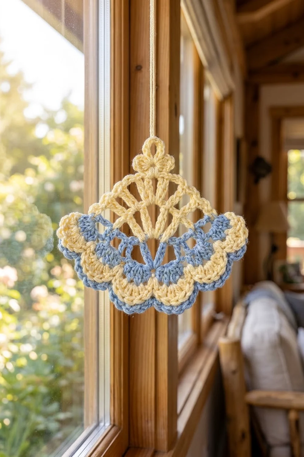 Cream and periwinkle crocheted flower ornament hanging in a bright wooden window frame with garden view beyond.
